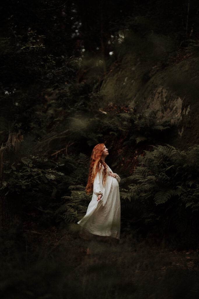 en gravid kvinna med långt rött hår dansar i skogen i en vit klänning