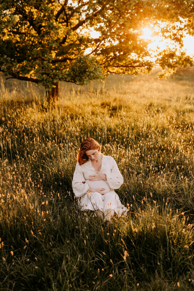 en gravid kvinna sitter i gräset med vackert guldigt ljus bakom henne