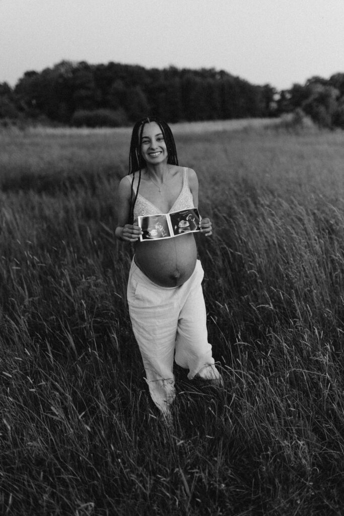 En gravid tvillingmamma står på ett fält och håller upp en ultraljudsbild som visar upp tvillingar