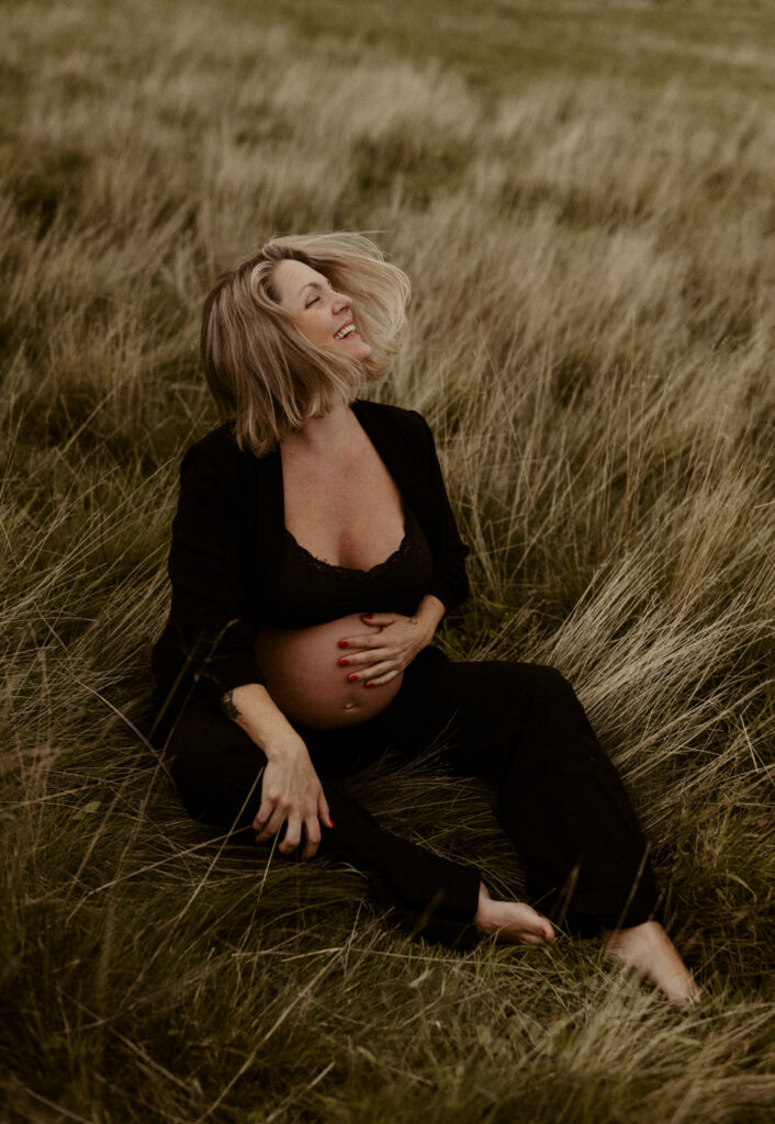 en mamma håller om sin gravidmage sittandes i gräset och skrattar