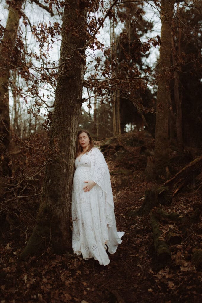 gravid kvinna i vit spetsklänning lutar sig mot ett träd i skogen