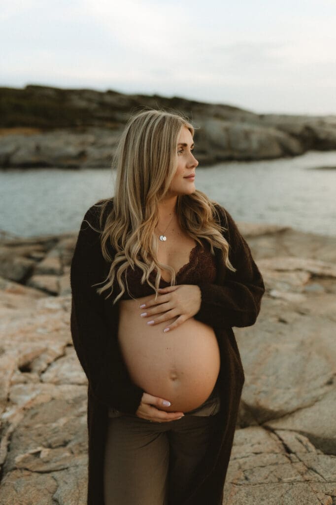 gravid kvinna i öppen brun kofta på klipporna vid havet, gravidfotografering på vintern i Göteborg