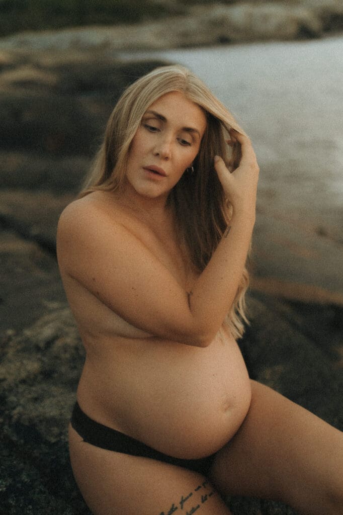 hudnära gravidbild utomhus där en gravid kvinna sitter på klipporna med handen i håret